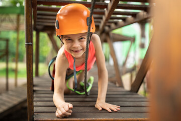 Portrait of little smiling girl in helmet and harness on trail in sky rope park in summer