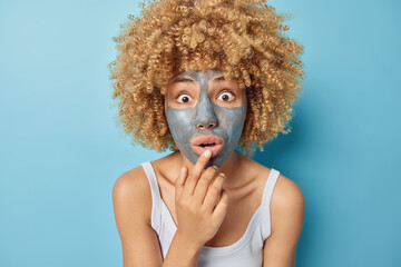 Portrait of shocked curly haired woman applies beauty clay mask on face for reducing pores keeps hand on lips dressed in casual white t shirt isolated over blue background. Wellness and spa.
