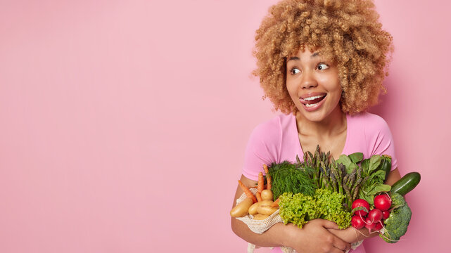 Cheerful surprised young woman with curly hair sticks out tongue looks wondered aside embraces different vegetables grown in own garden keeps to healthy nutrition isolated over pink background