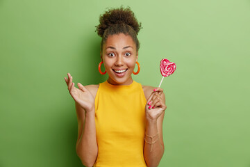 Photo of beautiful woman with dark curly hair smiles gladfully holds caramel lollipop dressed in yellow t shirt round earrings has happy mood isolated over green background. Sweet tooth concept