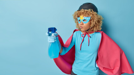 Serious curly woman wears eyemask helmet pink cloak and rubber gloves holds cleaning detergent washes dirty surface ready to help you with housework isolated over blue background blank space