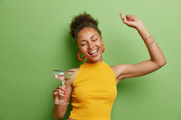 Horizontal shot of cheerful curly haired woman dances carefree wears yellow t shirt and earrings has pink manicure holds glass of cocktail isolated over vivid green background has fun on party.