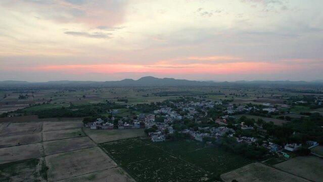 Height gain over square feilds and city in the distance with red sunset in the sky in front of mountains showing the rural village scenes in Indian outdoors and the beauty of the country