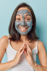 Glad young Asian woman with dark hair applies beauty mask undergoes anti aging procedures keeps hands together wants to have flawless skin isolated over blue background. Deep cleansing concept