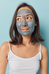 Pampering session after hard day. Dark haired woman with eastern appearance applies beauty mask to minimise pores or dark spots concentrated aside wears t shirt isolated over blue background