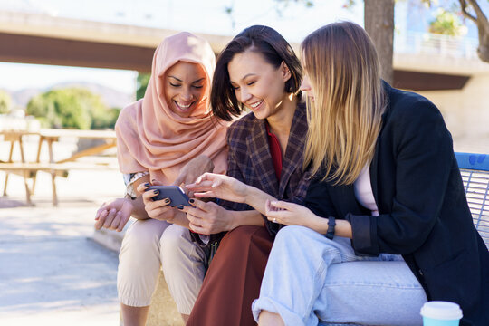 Positive multiethnic women sharing smartphone while resting on bench in park - Powered by Adobe