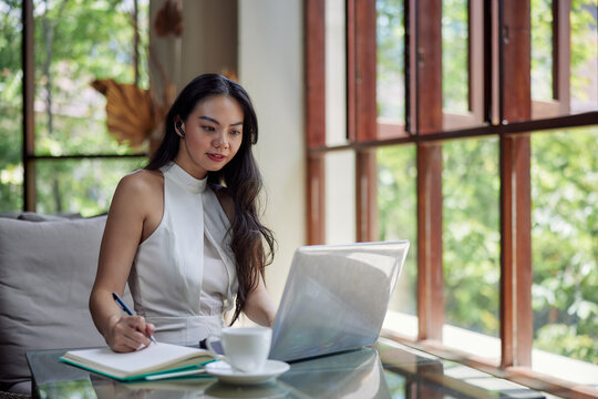 Young beautiful asian woman using laptop and writing notes, sitting in office, cafe or coworking space. Conference call, studying or working online
