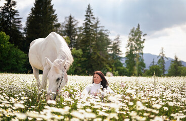 Woman sitting in a forest spring sun lit meadow full of daisy flowers, white Arabian horse next to...