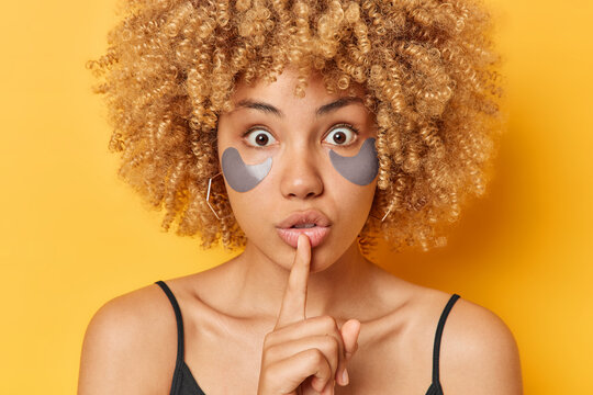 Photo of mysterious curly haired young woman makes silence gesture keeps inde finger over lips shares secret applies grey hydrogel patches under eyes stands against yellow background. Shh sign