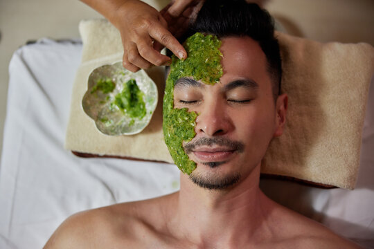 Spa therapist applying fresh cucumber mask on the face of young asian man. Male customer relaxing in beauty salon or spa
