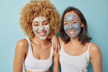 Home spa concept. Positive women with different appearance apply beauty clay mask on face for hydrating and moisturising skin dressed in white t shirts isolated over blue background. Skincare product