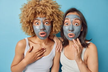 Indoor shot of stunned mixed race young women apply clay facial mask for skin treatment stare with shocked scared expressions dressed in t shirts isolated over blue background. Skin care concept