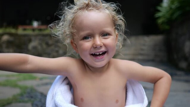 Cute baby in a towel smiles cheerfully. An adorable kid with an infectious laugh. A beautiful little girl of three years smiles sweetly and looks into the frame. Child blonde with curly hair.