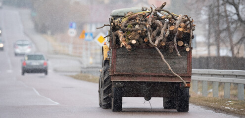 Tractor with a trailer loaded with wood on the road during snowfall. Transportation of firewood for...
