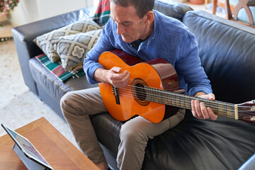 Mature man playing classical guitar with online course on tablet