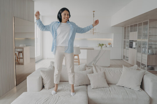 Excited Energetic Woman Wearing Headphones Having Fun Enjoying Music Jumping On Sofa Alone At Home