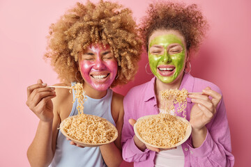 Cheerful women with curly hair applies beauty mask for skin treatment eat tasty spaghetti dressed in casual clothes stand next to each other isolated over pink background. Delicious noodles.