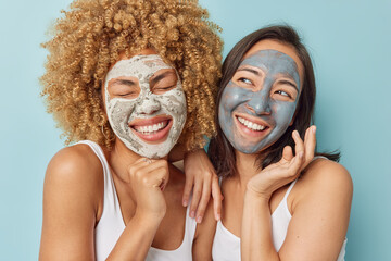 Photo of cheerful young friendly women smile happily apply beauty masks on face take care of facial skin have dreamy expressions isolated over blue background. Wellness and spa procedures concept