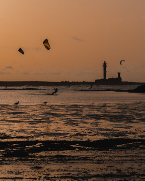 silhouettes of people kitesurfing on a paradisiacal beach with a lighthouse in the background in a beautiful sunset
