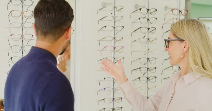 Optician helping a client choose glasses at an optometry appointment. Male customer in consult with optometrist fitting on a pair of eyewear spectacles for better eyesight and checking in the mirror