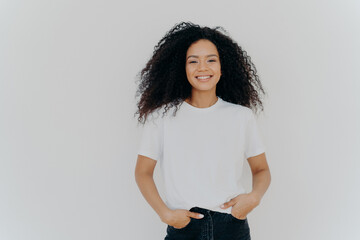 Isolated shot of young African American woman wears white t shirt, expresses good emotions, stands alone indoor, poses for photo, has casual talk with friend, enjoys free time. People and happiness