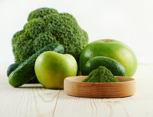 Green powder in wooden bowl on green background from vegetable and fruit. Chlorella or spirulina or matcha tea or dry spinach on wooden table. Concept dieting, detox, healthy superfood.
