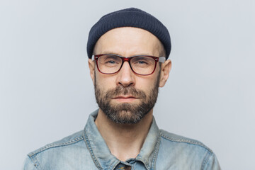 Photo of intelligent confident stylish man with dark thick beard and mustache, looks seriously into camera, poses against grey studio background, wears spectacles and hat. Facial expressions concept