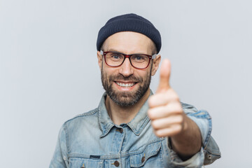 Satisfied attractive bearded male with happy expression, wears fashionable hat, denim jacket and glasses, keeps thumb raised, shows ok sign, demonstrates his approval, isolated on white wall