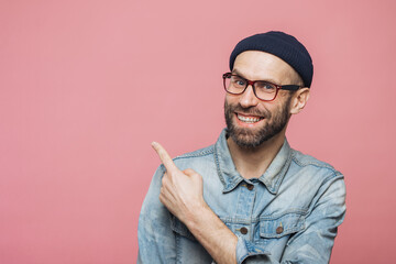 Advertisement concept. Cheerful young bearded man with glad expression wears eyewear, denim jacket...