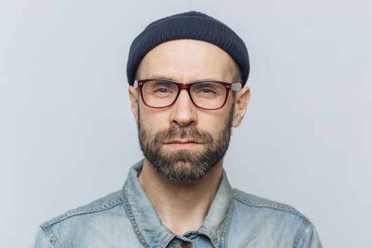 Photo of intelligent confident stylish man with dark thick beard and mustache, looks seriously into camera, poses against grey studio background, wears spectacles and hat. Facial expressions concept