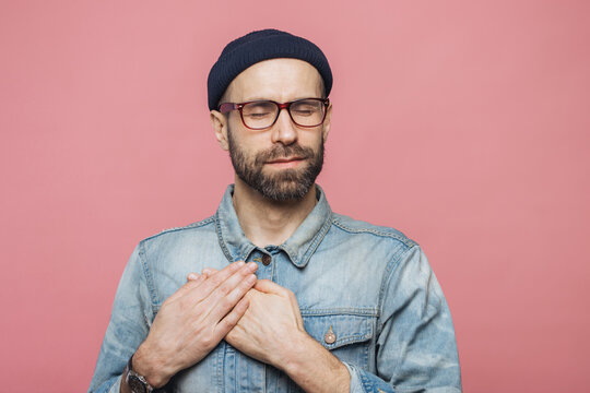 Pleased bearded man with closed eyes, keeps hands on chest, dressed in fashionable clothes, isolated over pink studio background. Middle aged male with shut eyes, expresses his love and sympathy