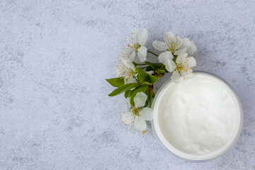Cosmetic facial body cream in glass jar and flowers on marble table. Minimalists still life with beauty products and white flowers on white background. Flat lay, top view, copy space