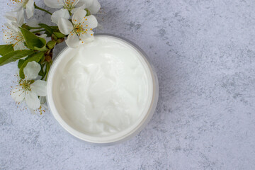 Cosmetic facial body cream in glass jar and flowers on marble table. Minimalists still life with beauty products and white flowers on white background. Flat lay, top view, copy space