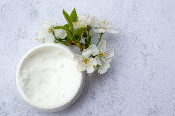 Fototapeta na wymiar Cosmetic facial body cream in glass jar and flowers on marble table. Minimalists still life with beauty products and white flowers on white background. Flat lay, top view, copy space