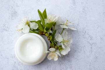Fototapeta na wymiar Cosmetic facial body cream in glass jar and flowers on marble table. Minimalists still life with beauty products and white flowers on white background. Flat lay, top view, copy space