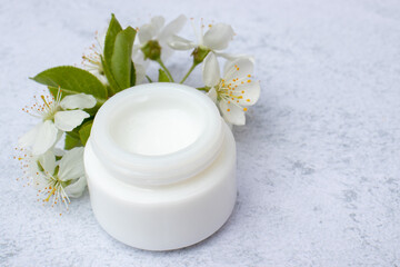 Fototapeta na wymiar Cosmetic facial body cream in glass jar and flowers on marble table. Minimalist still life with beauty products and white flowers on white background. FTop view, copy space