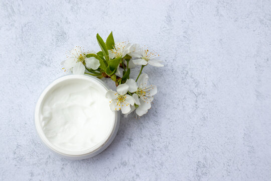 Cosmetic facial body cream in glass jar and flowers on marble table. Minimalists still life with beauty products and white flowers on white background. Flat lay, top view, copy space
