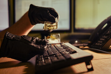 A man in black gloves with torn money dollars in his hands at a computer keyboard in a dark night office, close-up