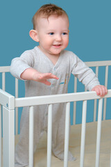 Happy infant baby boy stands in the crib, studio blue background. Smiling child in white pajamas