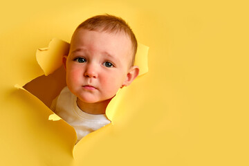 Sad child looks out of a hole in the studio yellow background. Cry baby boy peeks through a torn...