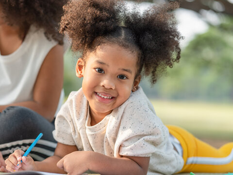 Smiling African American little girl with curly hair lying on mat drawing in the coloring book and looking at camera in the park.Kid girl learning and activity outdoor.