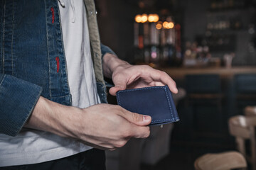 A young man stands in a cafe and holds a blue leather wallet in his hands. Lifestyle. Close-up, no face, horizontal orientation, copy space - obrazy, fototapety, plakaty