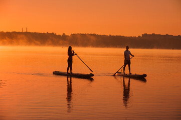 silhouettes of a couple on SUP boards on the water of the lake, against the background of the red sky with morning fog, active recreation