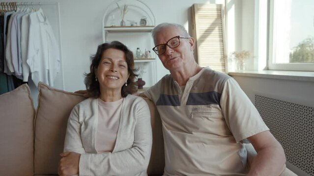 Cheerful elderly man and woman talk on camera saying goodbye and waving hands. Senior couple continues talking with son sitting on sofa from POV to third party