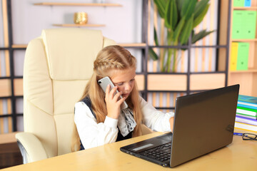 A young girl sits at a computer in the office. Schoolgirl in a laptop studies remotely. Office worker in an office with gadgets. Online training. Young teacher preparing for lessons