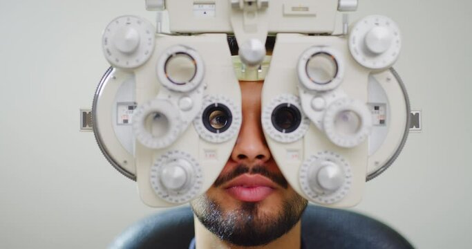 Male patient doing eye test with optometry machine. Closeup of a young man getting exam at an eyecare centre or ophthalmology clinic. Guy talking while looking through a phoropter to check his vision