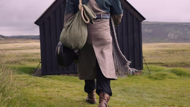 Stylish Man with a Sack Bag and a Banjo Case Walks Towards Little Black House