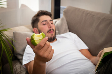 un jeune homme allongé sur un canapé se relaxe en mangeant une pomme et en lisant un livre