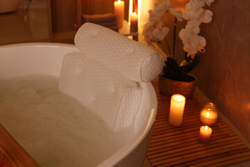 Tub with foamy water and soft bath pillow surrounded by candles indoors