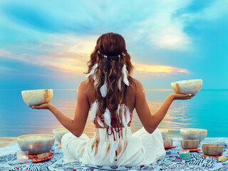 Woman with Tibetan Singing Bowls. Relaxation and Meditation at Sunset Beach. Sound Healing Therapy. Peaceful Women Silhouette practicing Yoga over Sunrise Sky - 517491777